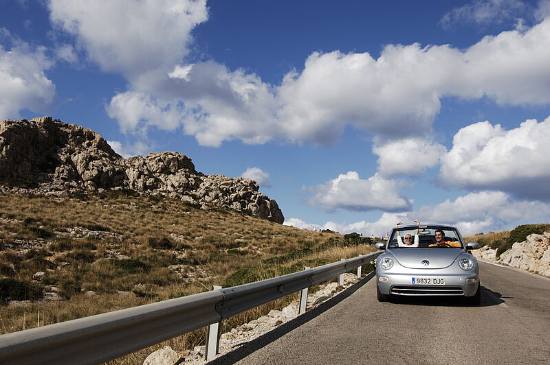 Silbernes Cabrio auf Bergstraße, felsige Landschaft und blauer Himmel mit weißen Wolken im Hintergrund.