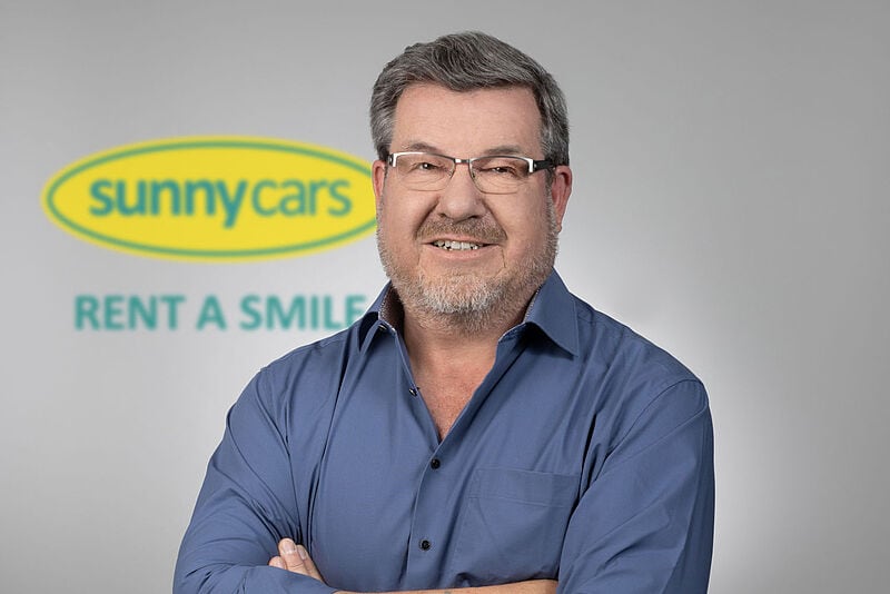 Professionelles Porträt eines Managers in blauem Hemd vor grauem Hintergrund mit dem Logo des Mietwagenvermittlers Sunny Cars und Slogan „Rent A Smile“.