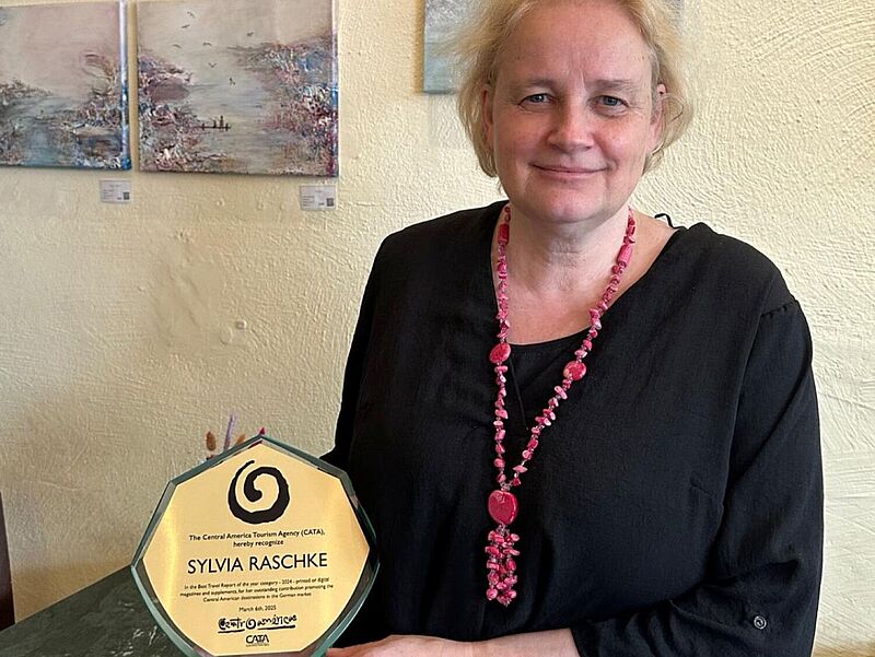 Eine Frau mit blondem Haar, schwarzer Bluse und pinkfarbener Halskette hält eine Auszeichnung aus Glas in der Hand. Die Auszeichnung trägt den Namen Sylvia Raschke und stammt von der Central America Tourism Agency (CATA). Im Hintergrund hängen mehrere Gemälde mit Meeres- und Küstenmotiven an einer hellen Wand.