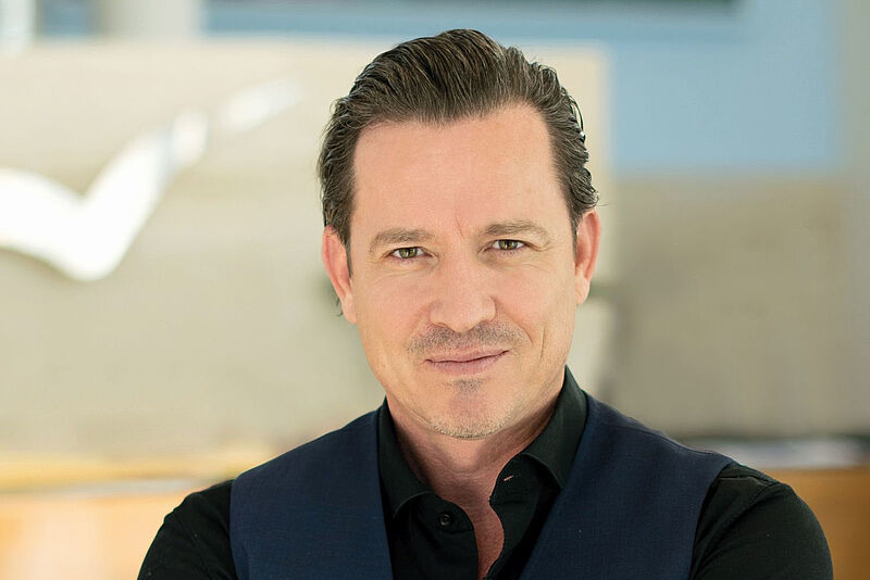 Ein Mann mittleren Alters mit kurzen, nach hinten gekämmten dunklen Haaren und einem leichten Bart steht mit verschränkten Armen und einem selbstbewussten Lächeln in einer modernen, hellen Umgebung. Er trägt ein schwarzes Hemd und eine dunkelblaue Weste. Im Hintergrund sind unscharfe architektonische Elemente und eine große, stilisierte Dekoration zu erkennen.