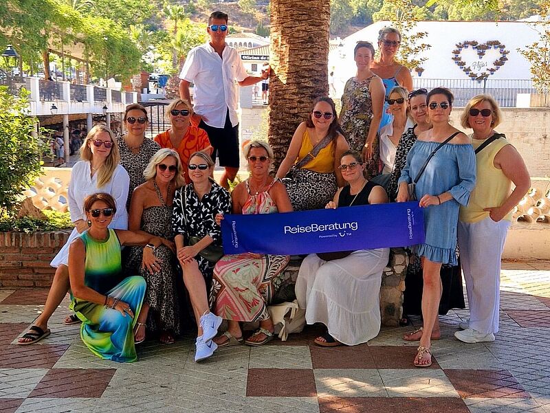 Gruppenfoto von 17 Personen, hauptsächlich Frauen, die unter einer großen Palme posieren. Die Gruppe gehört zur „Reiseberatung – powered by TUI“, was auf einem blauen Banner in der Mitte zu lesen ist. Im Hintergrund sind weiße Gebäude, Palmen und bewaldete Berge unter blauem Himmel zu sehen. Die Stimmung wirkt sonnig und entspannt – typisch für eine Reise in den Süden, vermutlich in Spanien.