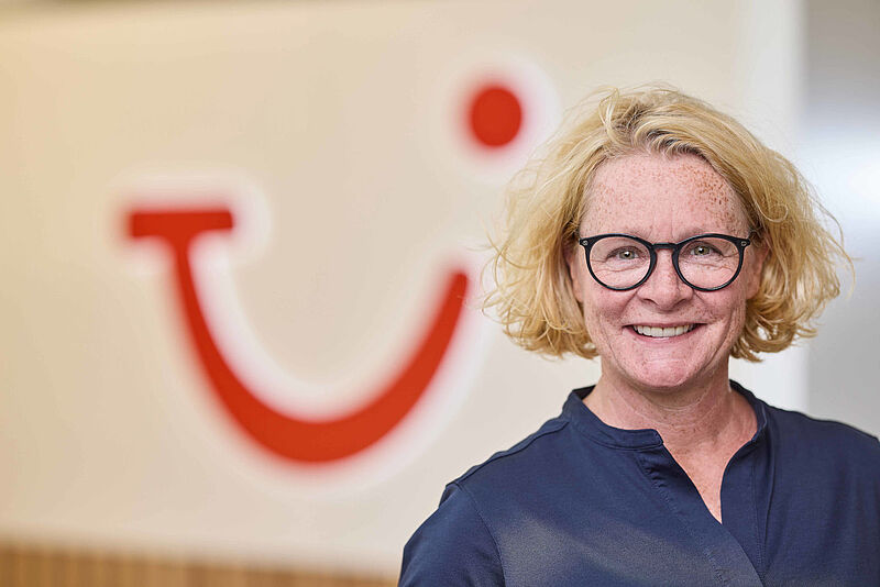 Ilka Lauenroth, Vertriebsdirektorin bei TUI, lächelt in die Kamera. Sie trägt eine dunkle Bluse und eine runde Brille, im Hintergrund ist unscharf das rote TUI-Logo erkennbar.