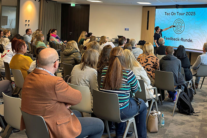 Ein moderner Konferenzraum mit einer Gruppe von Menschen, die an einer Präsentation teilnehmen. Ein Referent steht vorne und zeigt auf eine große Leinwand, auf der 'Tec On Tour 2025' und 'Feedback-Runde' zu lesen sind. Die Teilnehmenden sitzen in Reihen auf grauen Stühlen und hören aufmerksam zu. Viele tragen legere Kleidung, einige haben Jacken über die Stuhllehnen gehängt. Der Raum ist hell beleuchtet, mit einer Mischung aus natürlichen und künstlichen Lichtquellen