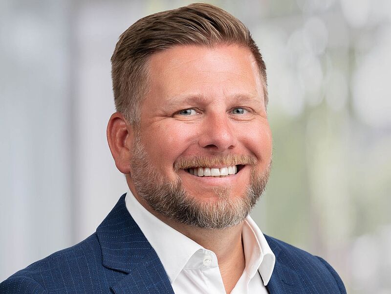 Ein lächelnder Mann mit kurzem, hellbraunem Haar und Bart trägt ein dunkelblaues Sakko über einem weißen Hemd. Der Hintergrund ist unscharf und hell, vermutlich in einem Büro oder Konferenzraum aufgenommen.