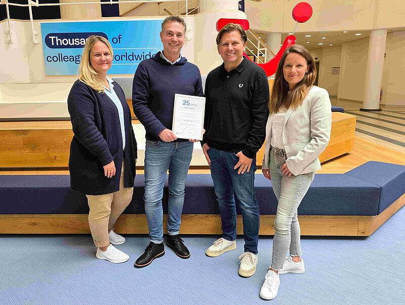 Vier Menschen, zwei Frauen und zwei Männer, lächeln in die Kamera. Ein Mann hält eine Urkunde in der Hand, auf der eine 25 zu sehen ist.