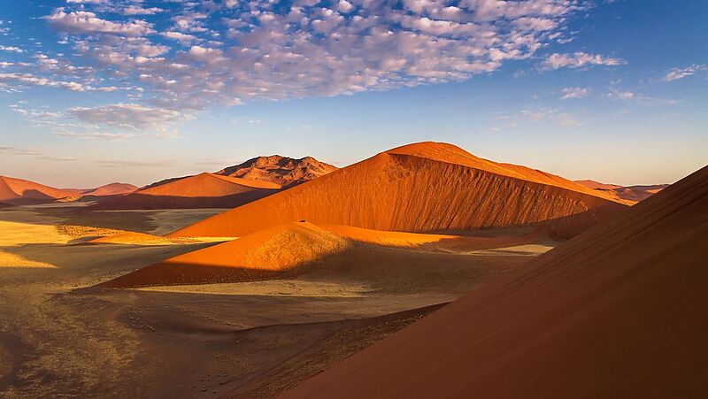 Sonnenbeschienene Sanddünen in warmen Orange- und Rottönen in einer Wüstenlandschaft, mit klarem blauem Himmel und vereinzelten weißen Wolken darüber.