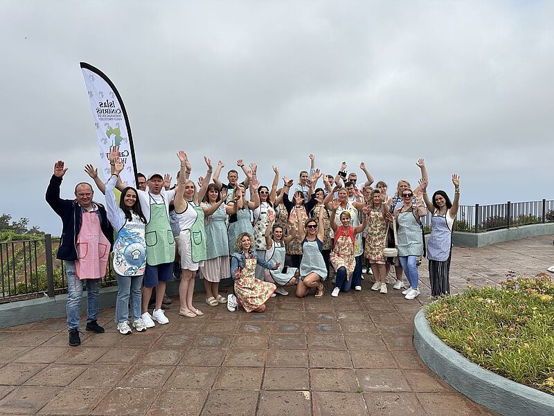 Eine große Gruppe fröhlicher Menschen steht im Freien auf einer Terrasse mit Meerblick, viele tragen bunte Schürzen und heben die Arme zum Gruß. Im Hintergrund ist ein Werbeflagge mit der Aufschrift 