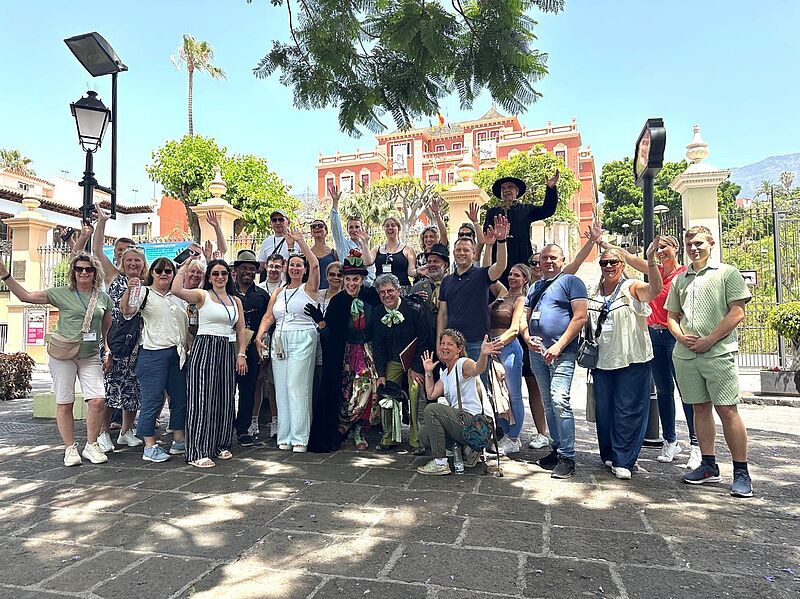 Eine fröhliche Gruppe von etwa 30 Personen steht im Freien vor einer historischen Kulisse mit bunten Gebäuden, Palmen und dekorativem Tor. Die Menschen lachen, winken und posieren für das Gruppenfoto. Zwei Personen in der Mitte sind kostümiert, eine davon trägt einen schwarzen Hut und lange schwarze Kleidung, die andere ein farbenfrohes Outfit mit grünem Halstuch. Im Hintergrund ist ein rotes Gebäude mit weißem Stuck sichtbar, vermutlich in mediterranem Stil. Das Wetter ist sonnig und klar.