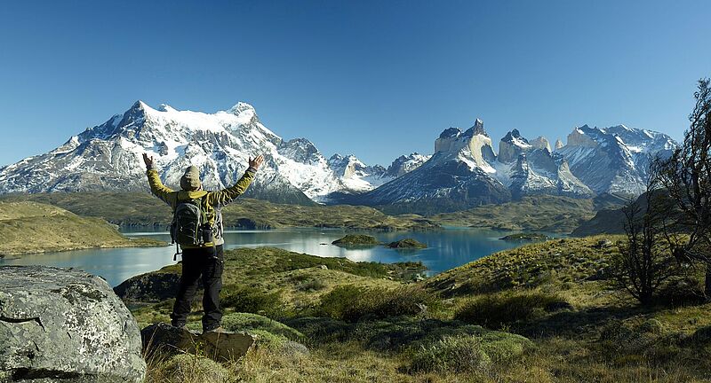 Ein Wanderer mit grünem Rucksack und gelber Jacke steht auf einem Felsen und streckt triumphierend die Arme in die Höhe. Vor ihm erstreckt sich die atemberaubende Landschaft des Torres del Paine Nationalparks in Chile mit schneebedeckten Gipfeln, tiefblauen Seen und weitläufigen Ebenen unter einem klaren, strahlend blauen Himmel. Links im Vordergrund ein großer Felsblock, rechts einige kahle Bäume.