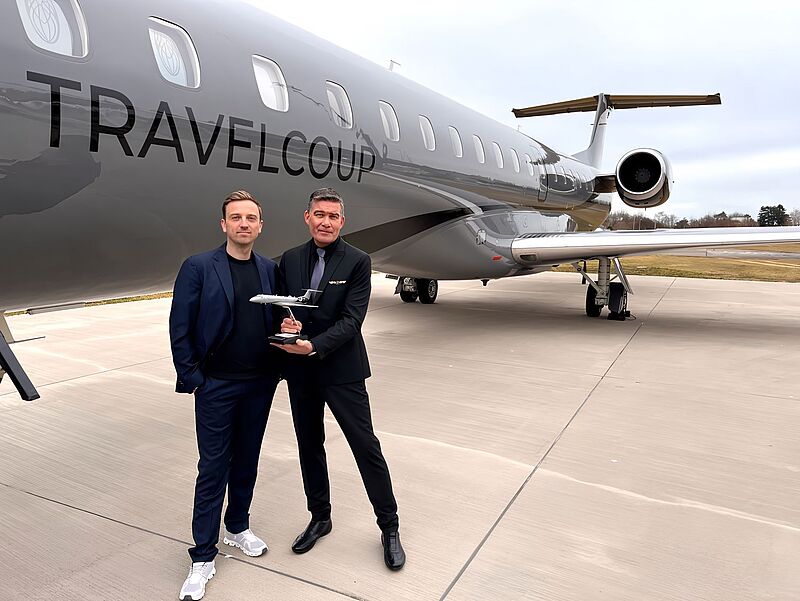 Zwei Männer stehen vor einem luxuriösen Privatjet mit der Aufschrift 'TRAVELCOUP' auf dem Rumpf. Der Mann links trägt einen dunklen Anzug mit einem schwarzen Shirt und weißen Sneakern, während der Mann rechts einen schwarzen Anzug mit blauer Krawatte trägt und ein Modellflugzeug auf einem Sockel hält. Der Hintergrund zeigt eine Rollbahn mit etwas Grasfläche und Bäumen unter einem bewölkten Himmel