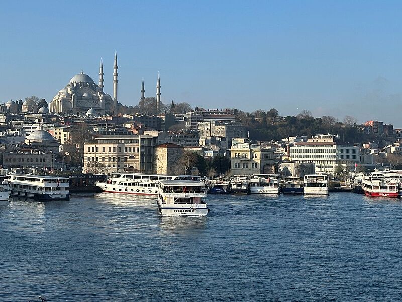 Blick über das Wasser des Goldenen Horns in Istanbul, Türkei, mit zahlreichen weißen und bunten Ausflugsbooten im Vordergrund. Im Hintergrund erhebt sich die imposante Süleymaniye-Moschee mit ihren hohen Minaretten über die dicht bebaute Altstadt. Der Himmel ist klar und blau, die Szene wirkt ruhig und sonnig.