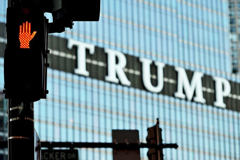 Rote LED-Hand eines Fußgängerampelsignals vor verschwommenem Trump-Schriftzug an Glasfassade eines Hochhauses.