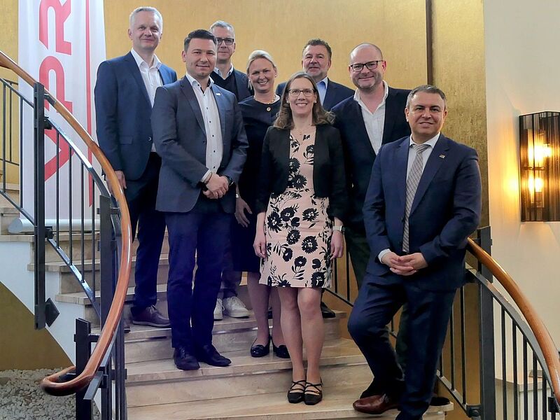 Acht Personen stehen auf einer eleganten Steintreppe in einem Innenraum mit goldfarbener Wand. Es handelt sich um sechs Männer und zwei Frauen in Businesskleidung, die für ein Gruppenfoto posieren. Im Hintergrund steht ein weißes Banner mit rotem „VPR“-Logo. Die Atmosphäre wirkt professionell, das Bild wurde vermutlich im Rahmen eines offiziellen Anlasses oder einer Veranstaltung aufgenommen. Alle Personen lächeln in die Kamera. Auf der untersten Stufe ist ebenfalls das rote VPR-Logo aufgebracht.