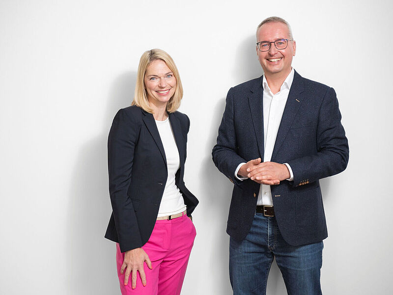 Eine Managerin und ein Manager in legerer Business-Kleidung stehen lächelnd vor einer weißen Wand.