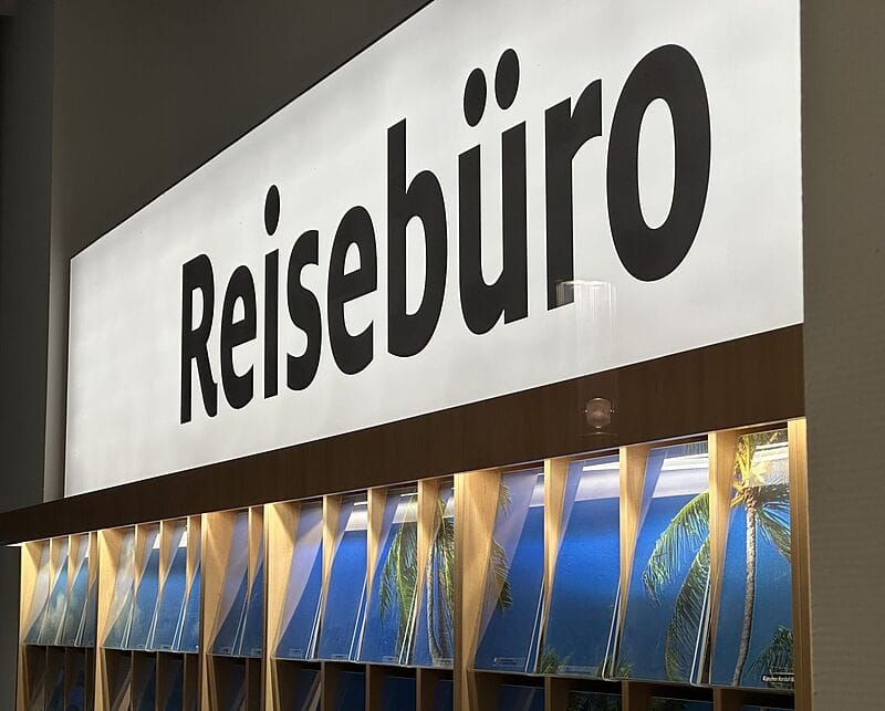 Seitlicher Blick auf ein weißes Leuchtboard, auf dem in schwarzen Buchstaben das Wort Reisebüro steht.