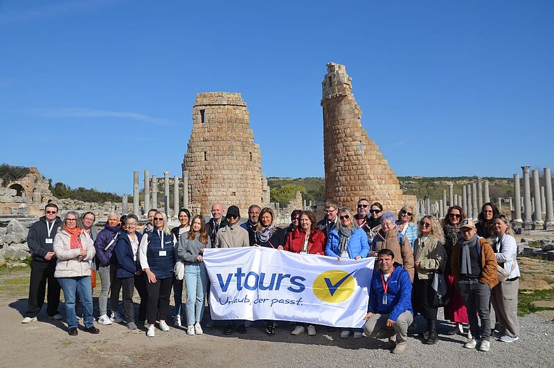 Bei strahlendem Sonnenschein präsentieren sich 20 Personen in der türkischen Ausgrabungsstätte Perge, sie halten eine Fahne des Reiseveranstalters Vtours