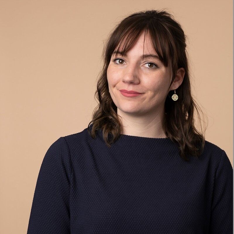 Portrait einer jungen Frau mit schulterlangem, braunem, gewelltem Haar und Pony. Sie trägt ein dunkelblaues, strukturiertes Oberteil und goldene Ohrringe mit rundem Anhänger. Der Hintergrund ist in einem einfarbigen, warmen Beigeton gehalten. Die Frau blickt freundlich in die Kamera und lächelt leicht.