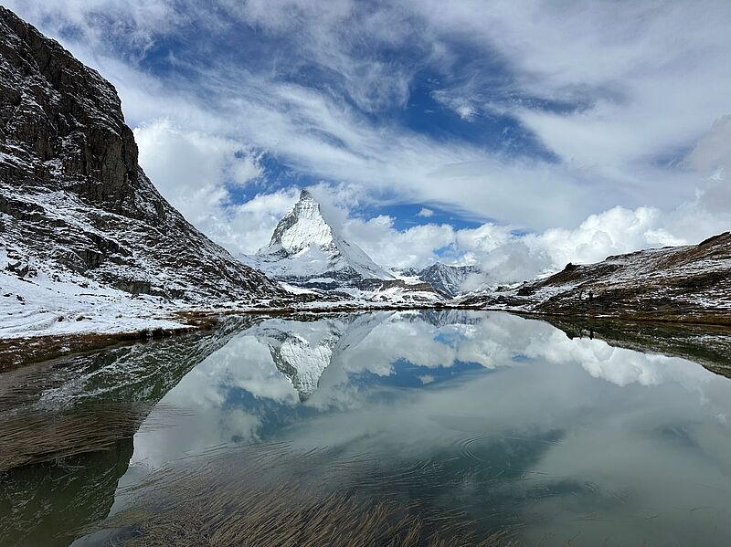 Schneebedeckte Berglandschaft mit dem markanten Matterhorn im Hintergrund, das sich klar in einem ruhigen Bergsee spiegelt. Wolken und blauer Himmel verstärken die dramatische Atmosphäre, während die felsigen Hänge und Schneeflächen den Vordergrund prägen.
