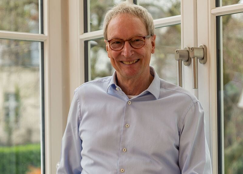 Ein älterer Mann mit grauen Haaren und Brille steht lächelnd vor einem Fenster. Er trägt ein hellblaues Hemd und lehnt sich leicht zur Seite. Im Hintergrund sind Bäume und ein Gebäude durch die Fensterscheiben sichtbar.