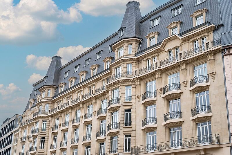 Fassade eines eleganten historischen Gebäudes mit französischen Balkonen und reich verzierten Fenstern unter einem Mansardendach, aufgenommen bei Tageslicht unter blauem Himmel mit vereinzelten Wolken