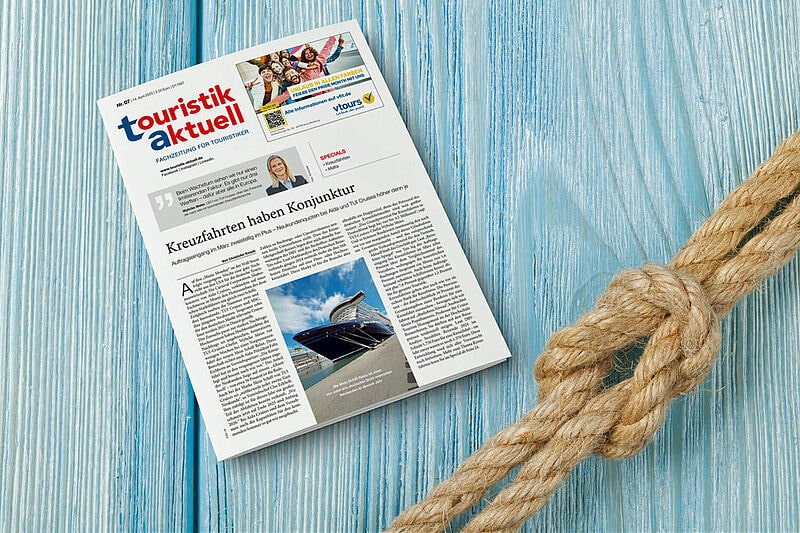 Eine Ausgabe der Fachzeitung „touristik aktuell“ liegt auf einem hellblauen, hölzernen Untergrund mit maritimer Atmosphäre. Daneben ist ein dicker, geknoteter Tau zu sehen. Auf der Titelseite der Zeitung ist ein Artikel mit dem Titel „Kreuzfahrten haben Konjunktur“ sowie ein Bild eines Kreuzfahrtschiffs abgebildet.
