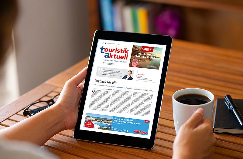 Eine Person hält ein Tablet in der linken Hand, auf dem die digitale Ausgabe der Fachzeitung touristik aktuell angezeigt wird. In der rechten Hand hält die Person eine weiße Kaffeetasse. Auf dem Tablet ist ein Artikel mit dem Titel „Payback für alle“ zu sehen sowie Werbeanzeigen für Reiseangebote. Im Hintergrund liegt eine Brille auf dem Holztisch, daneben ein Notizbuch mit einem Stift.