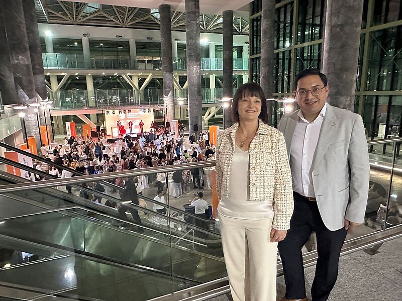 Zwei Personen in eleganter Kleidung stehen auf einer Galerie mit Glasgeländer in einem modernen, hell erleuchteten Gebäude. Im Hintergrund ist eine Veranstaltung mit vielen Gästen, Tischen und einer Bühne mit Künstlern zu sehen.