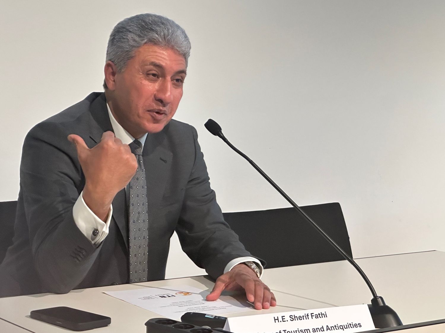 Mann im grauen Anzug sitzt auf Podium vor Mikrofon