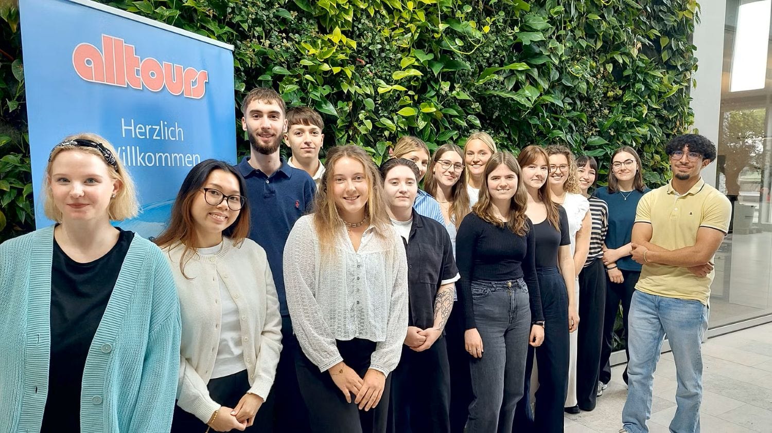 Gruppe junger Auszubildender steht lächelnd vor dem Alltours-Firmenschild, einige halten Ordner oder Taschen, sonniges Wetter.