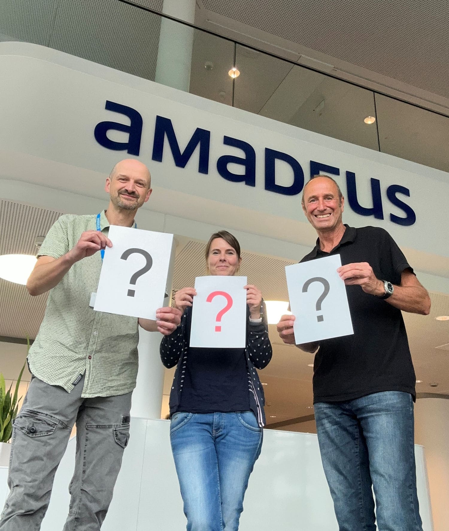Drei Personen stehen vor einem Schild mit der Aufschrift ‚Amadeus‘ und halten jeweils ein Blatt Papier mit einem großen Fragezeichen in den Händen. Die mittlere Person hält ein rotes Fragezeichen, die beiden äußeren schwarze.