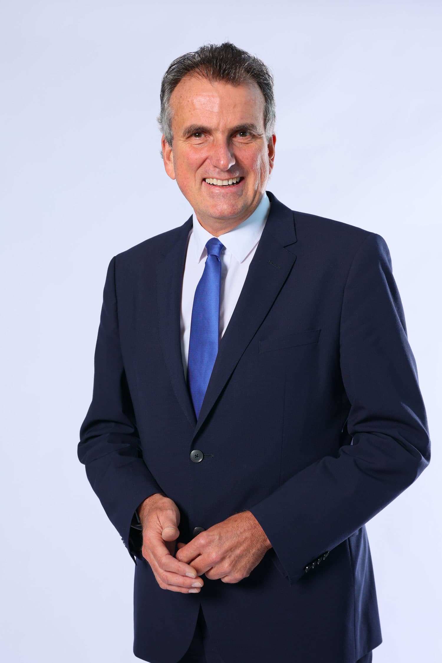 Vor einer weißen Fotolandwand posiert ein lächelnder Reederei-Manager mit braunem Haar und grauem Haaransatz. Er trägt einen dunklen Anzug mit blauer Krawatte auf weißem Oberhemd.