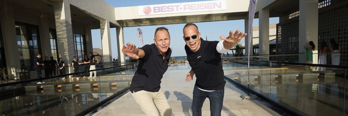 Zwei Männer mittleren Alters im Freizeitlook zeigen sich dynamisch vor dem Eingang eines Tagungshotels.