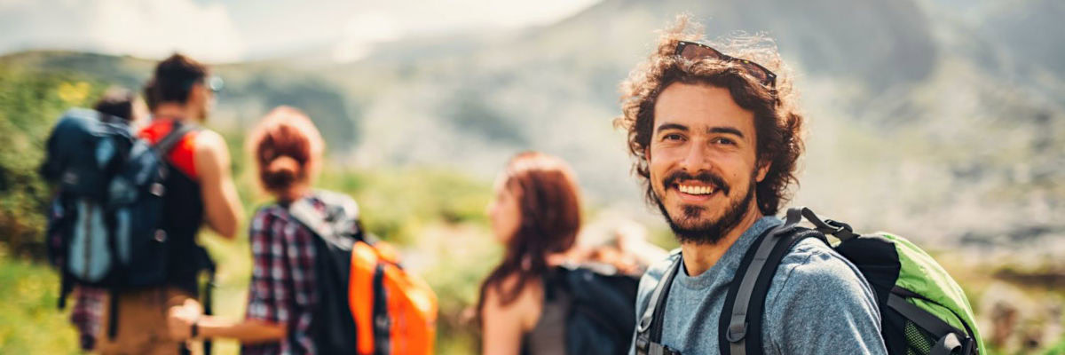 Eine Gruppe von Wandernden auf einem Bergpfad; im Vordergrund steht ein junger Mann mit Rucksack und Trekkingstöcken, der lächelnd in die Kamera blickt, während die übrigen Personen leicht unscharf im Hintergrund weitergehen.