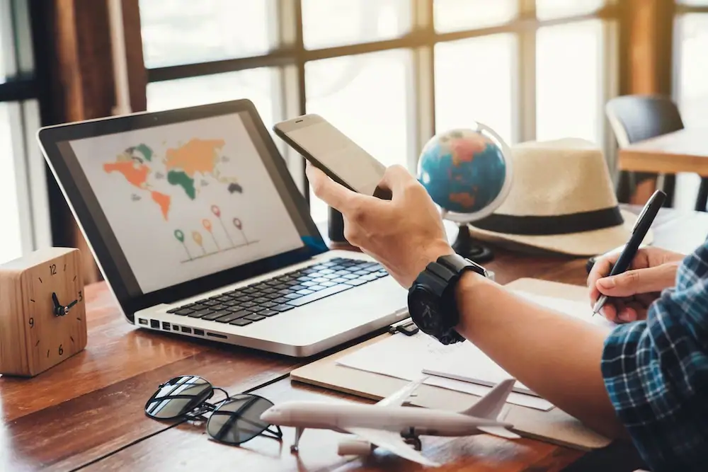 Schreibtischsituation in einem Reisebüro oder Homeoffice: Eine Person hält ein Smartphone in der Hand und schreibt gleichzeitig auf einem Notizblock. Vor ihr stehen ein Laptop mit einer Weltkarte auf dem Bildschirm, eine kleine Globusfigur, eine Sonnenbrille, ein Modellflugzeug und ein Strohhut.