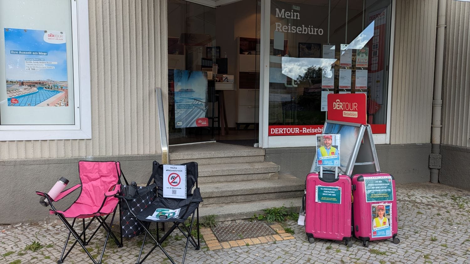 Zwei leere Campingstühle mit Streikschildern und Informationsmaterial stehen vor dem Eingang eines Dertour-Reisebüros. Daneben zwei pinke Rollkoffer mit ver.di-Plakaten und einem Schild „Heute Reisestreik“. Ein Aufsteller mit weiteren Informationen zur Streikaktion ergänzt das Bild. Das Reisebüro ist geschlossen, im Fenster hängt Werbung für Urlaubsangebote.