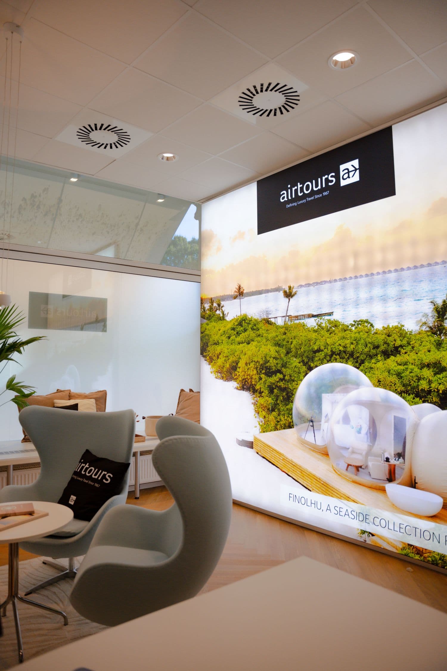 Beratungslounge im Airtours-Design im TUI-Flagshipstore Aschaffenburg mit hellen Sesseln, Dekokissen und großem Wandbild einer tropischen Inselkulisse.
