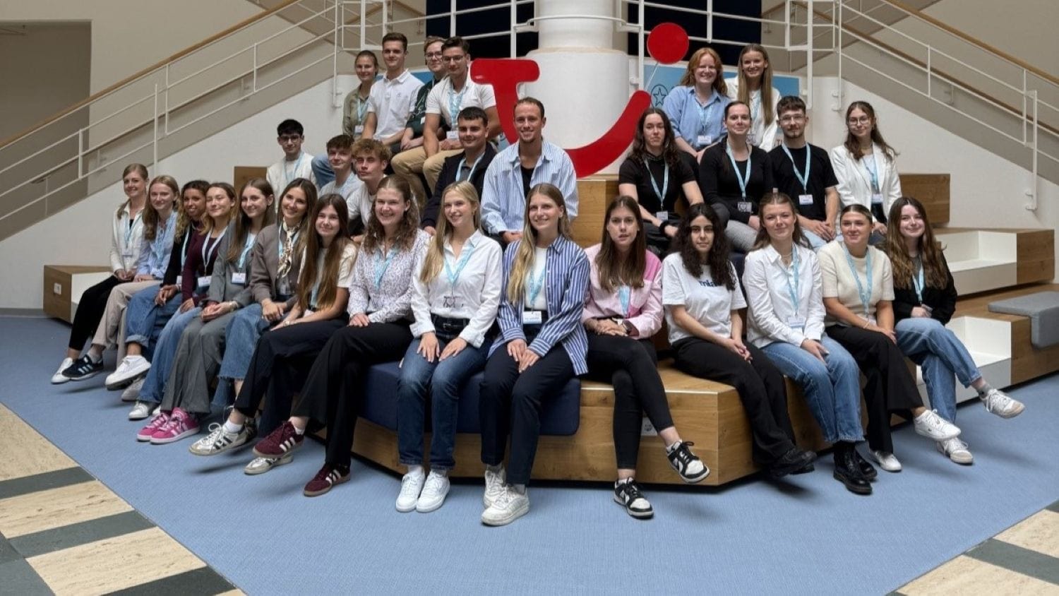 Große Gruppe neuer Auszubildender sitzt und steht auf Holzstufen im TUI-Campus vor einem großen roten TUI-Logo; alle tragen Namensschilder und hellblaue Schlüsselbänder, lächelnd in die Kamera blickend.
