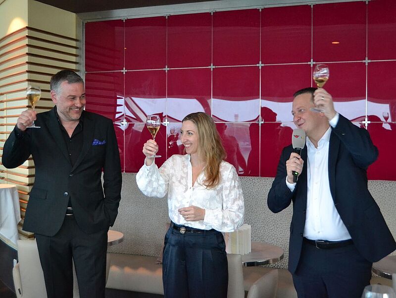 In der Lounge eines Aida-Kreuzfahrtschiffes prosten sich bei einem Geschäftsempfang drei strahlende Verantwortliche der Reederei mit gefüllten Sektgläsern zu.