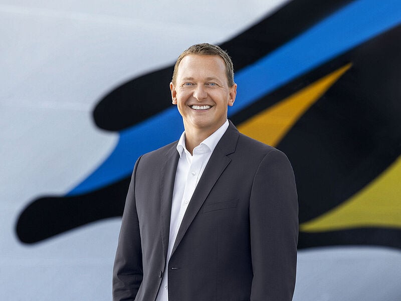 Ein Reederei-Manager Mitte 40 mit kurzen braunen Haaren und blauen Augen steht vor einem Kreuzfahrtschiff und blickt freudig lächelnd in die Kamera.