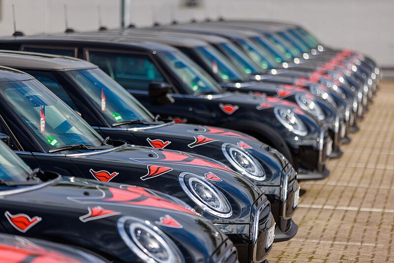 Schwarze Autos der Marke Mini Cooper stehen in einer Reihe, versehen mit roten Logos der Kreuzfahrtreederei Aida. Sie stehen nebeneinander geparkt auf gepflastertem Untergrund, die vorderen Autos sind scharf, die hinteren unscharf.