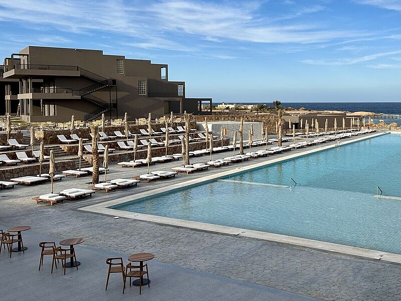 Luxuriöse Poolanlage eines Resorts mit zahlreichen weißen Sonnenliegen und geschlossenen Sonnenschirmen entlang des Wassers. Im Hintergrund befindet sich ein modernes, dunkel gehaltenes Gebäude mit Außentreppen sowie das blaue Meer mit einem Steg. Der Himmel ist klar und sonnig. Die Szenerie strahlt Ruhe und Exklusivität aus.