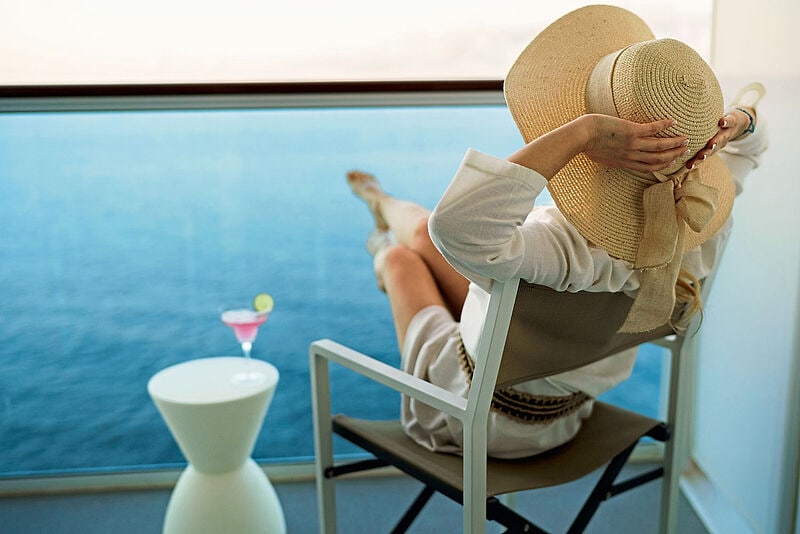 Eine allein reisende Frau sitzt auf dem Balkon eines Kreuzfahrtschiff, blickt aufs Meer hinaus und streckt genussvoll die Arme von sich.