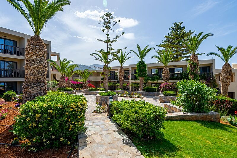 Gepflegte Hotelanlage mit modernem, flachem Gebäude im mediterranen Stil, umgeben von Palmen, blühenden Sträuchern und gepflegten Steinwegen bei sonnigem Wetter auf Kreta