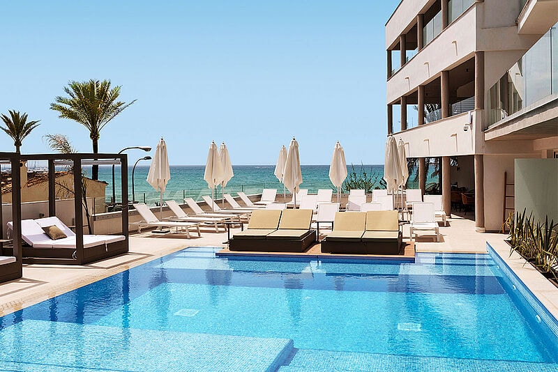 Infinity-Pool mit Meerblick, weißen Liegestühlen und Sonnenschirmen, umgeben von Palmen vor moderner Hotelarchitektur.