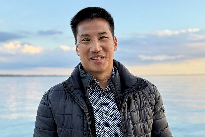 Mann in Winterjacke und Hemd lächelt vor Meereshorizont bei Sonnenuntergang, blauer Himmel mit Wolken im Hintergrund.