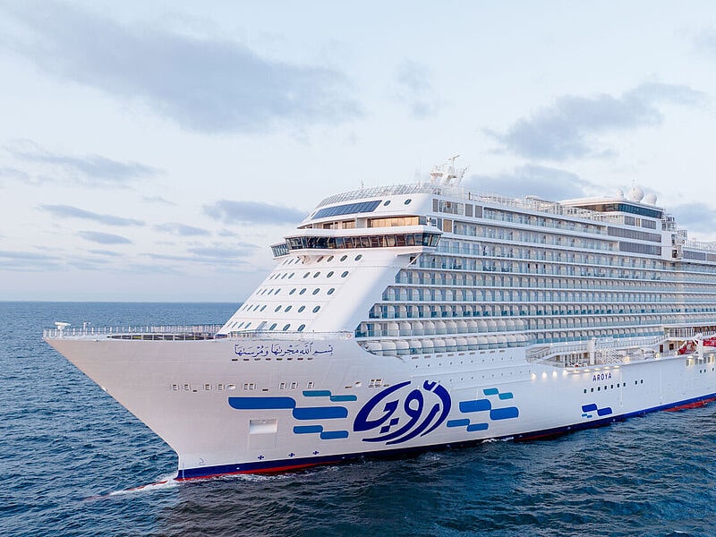 Ein neues saudisches Kreuzfahrtschiff mit weißem Raumpf und Aufbau fährt auf blauem Meer. Am Rumpf sind aufgehängte rot-weiße Rettungsboote und aufgemalte blaue Rechtecke zu sehen.