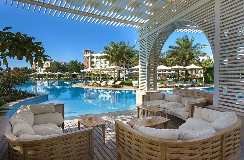 Luxuriöse Poolanlage mit weißer Pergola-Lounge, Liegestühlen, Sonnenschirmen und Palmen vor mediterraner Hotelarchitektur mit Turm.