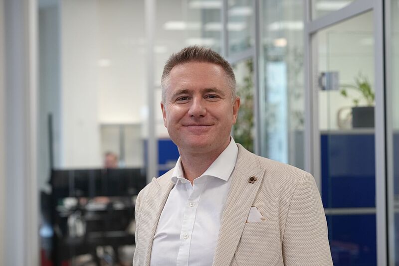 Ein Mann mittleren Alters im hellen Sakko und weißem Hemd steht in einem modernen Büroflur, lächelt freundlich in die Kamera.