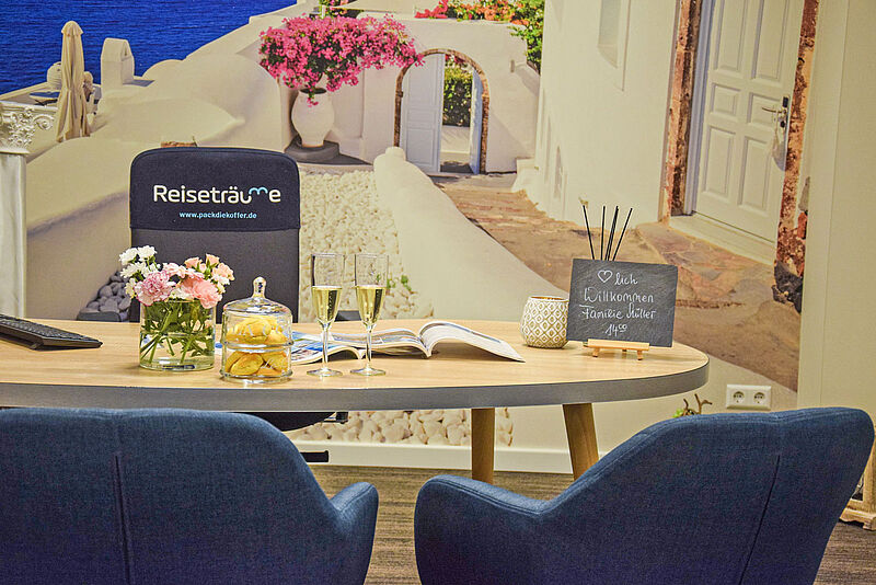 Ein einladender Beratungstisch in einem Reisebüro, dekoriert mit frischen Blumen, Gebäck unter einer Glasglocke, zwei Gläsern Sekt und einem offenen Reisekatalog. Auf einem kleinen Schild steht „Herzlich Willkommen Familie Müller 14:00“. Im Hintergrund ist eine Wand mit einem Urlaubs-Motiv von weißen Häusern und pinken Bougainvillea-Blüten im mediterranen Stil zu sehen.
