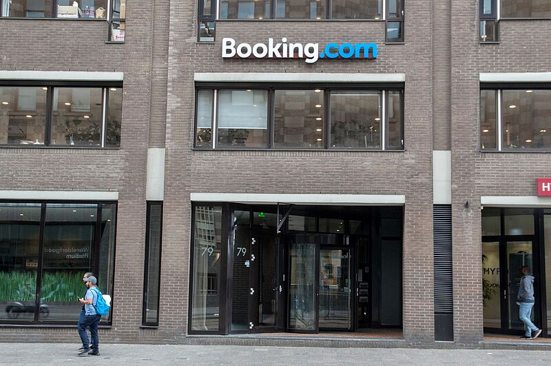 Ein Mann in blauem T-Shirt und Jeans geht an einem Backsteingebäude vorbei, auf dem groß Booking steht